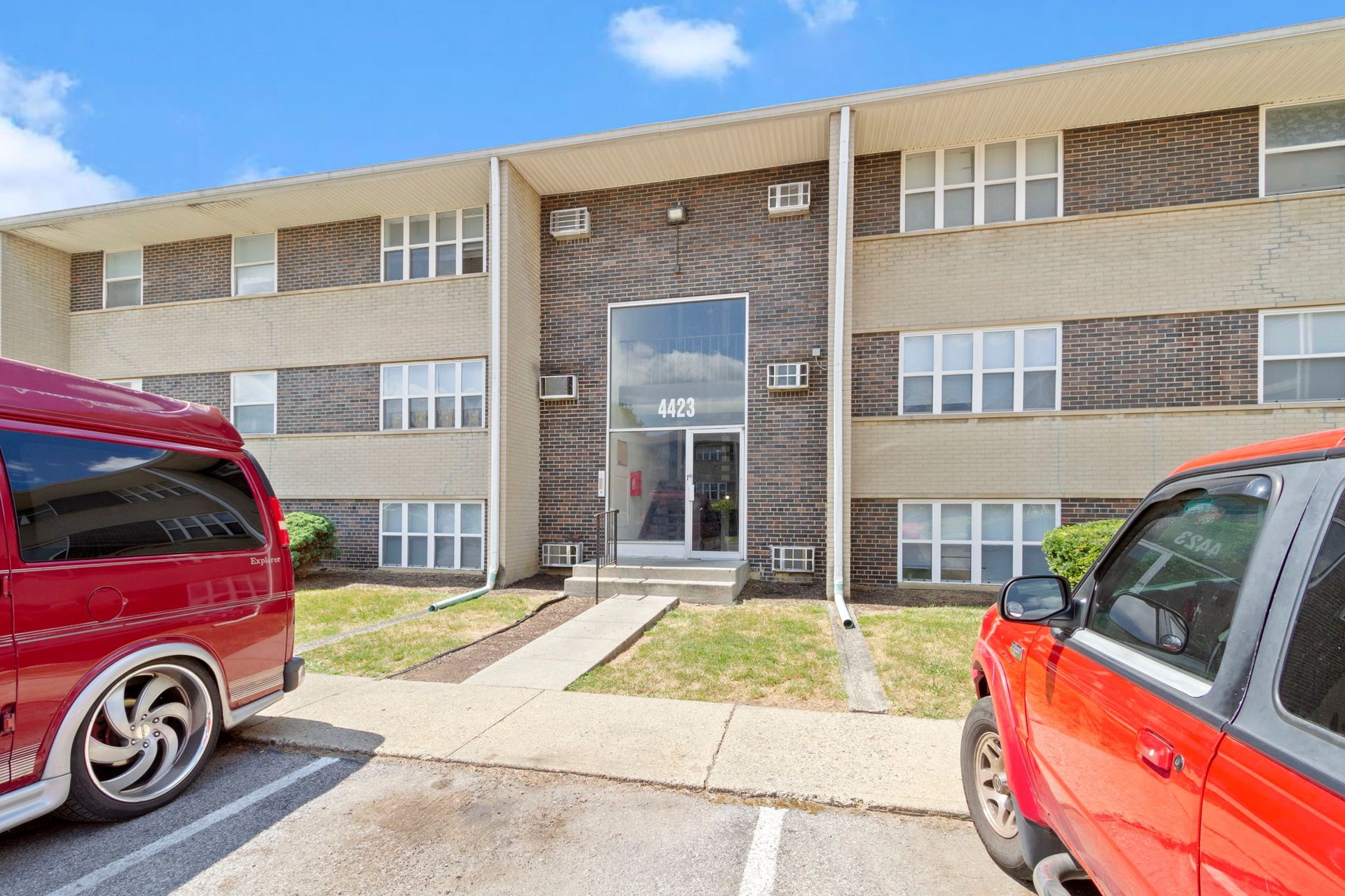 Apartment building with red van and truck in front, blue sky.