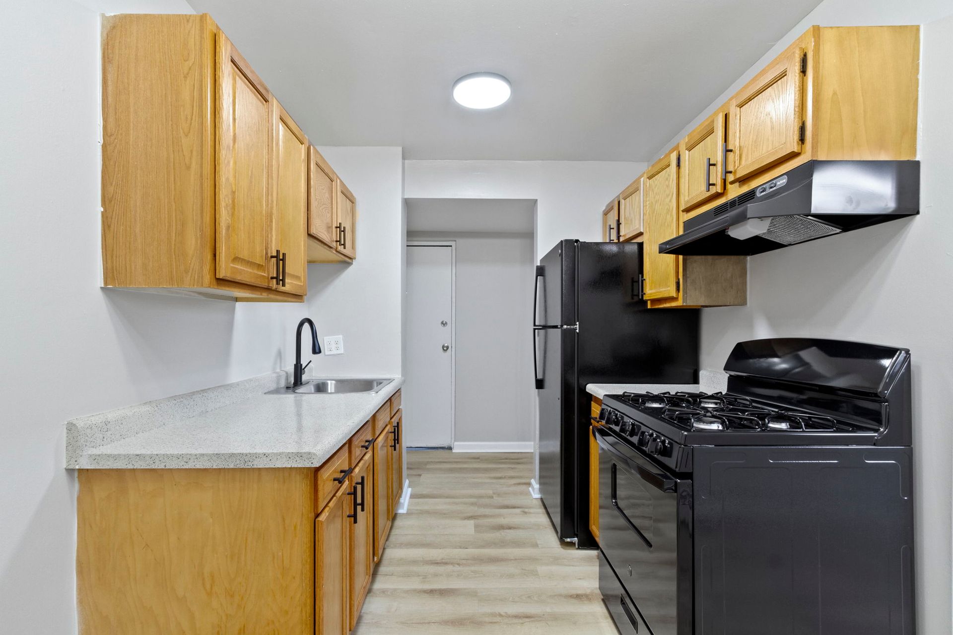 Narrow kitchen with light wood cabinets, black appliances, and light countertops.