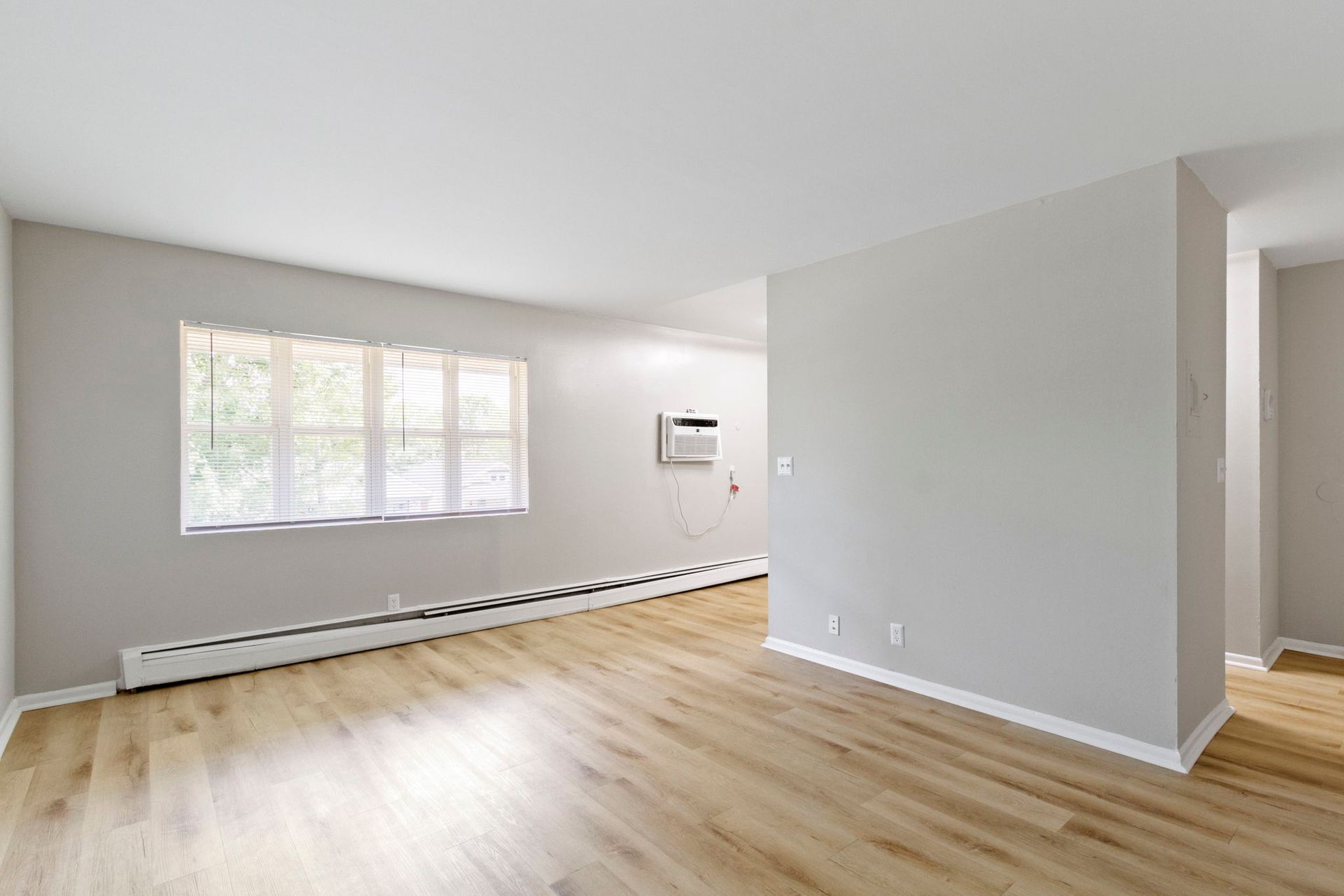 Empty living room with light-colored walls, wood-look flooring, a window, and an AC unit.