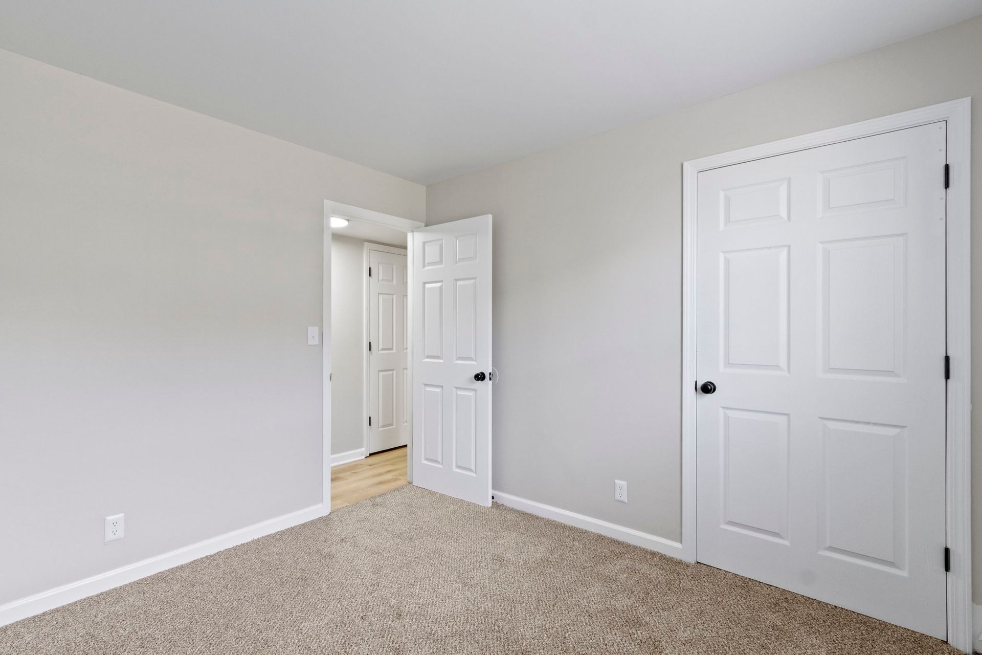 Empty bedroom with beige carpet, white walls and doors, and a doorway to another room.