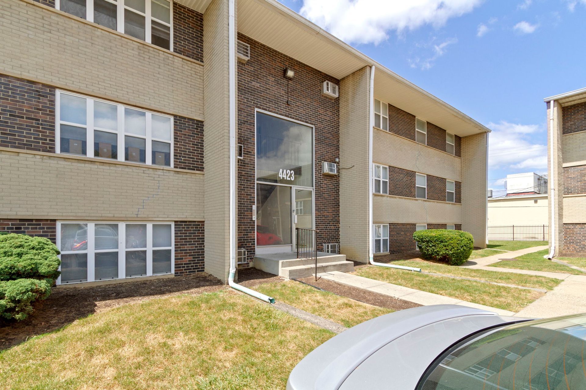 Apartment building exterior with tan brick, glass entry, and patches of grass and car.