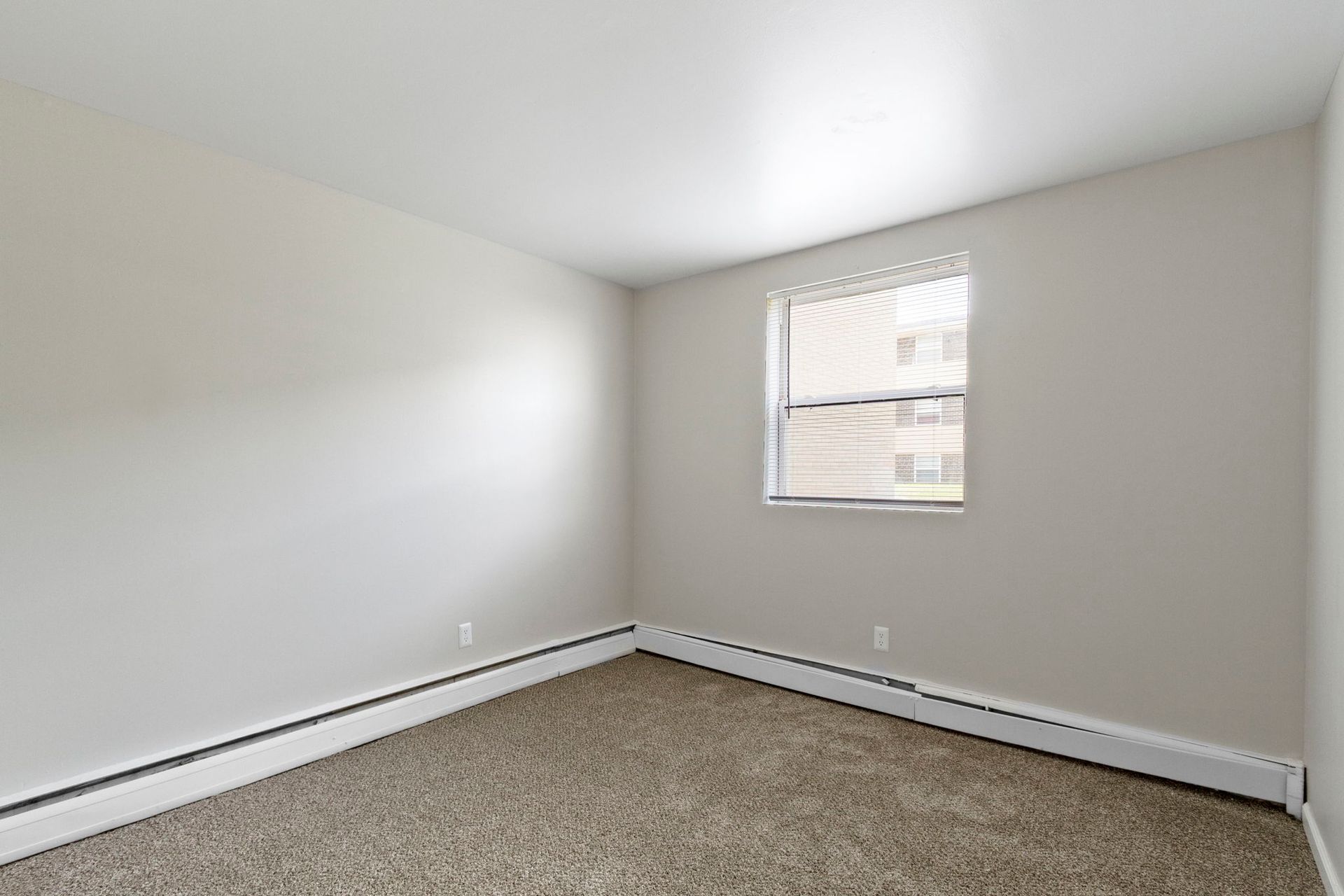 Empty bedroom with beige walls, brown carpet, and a window with blinds.