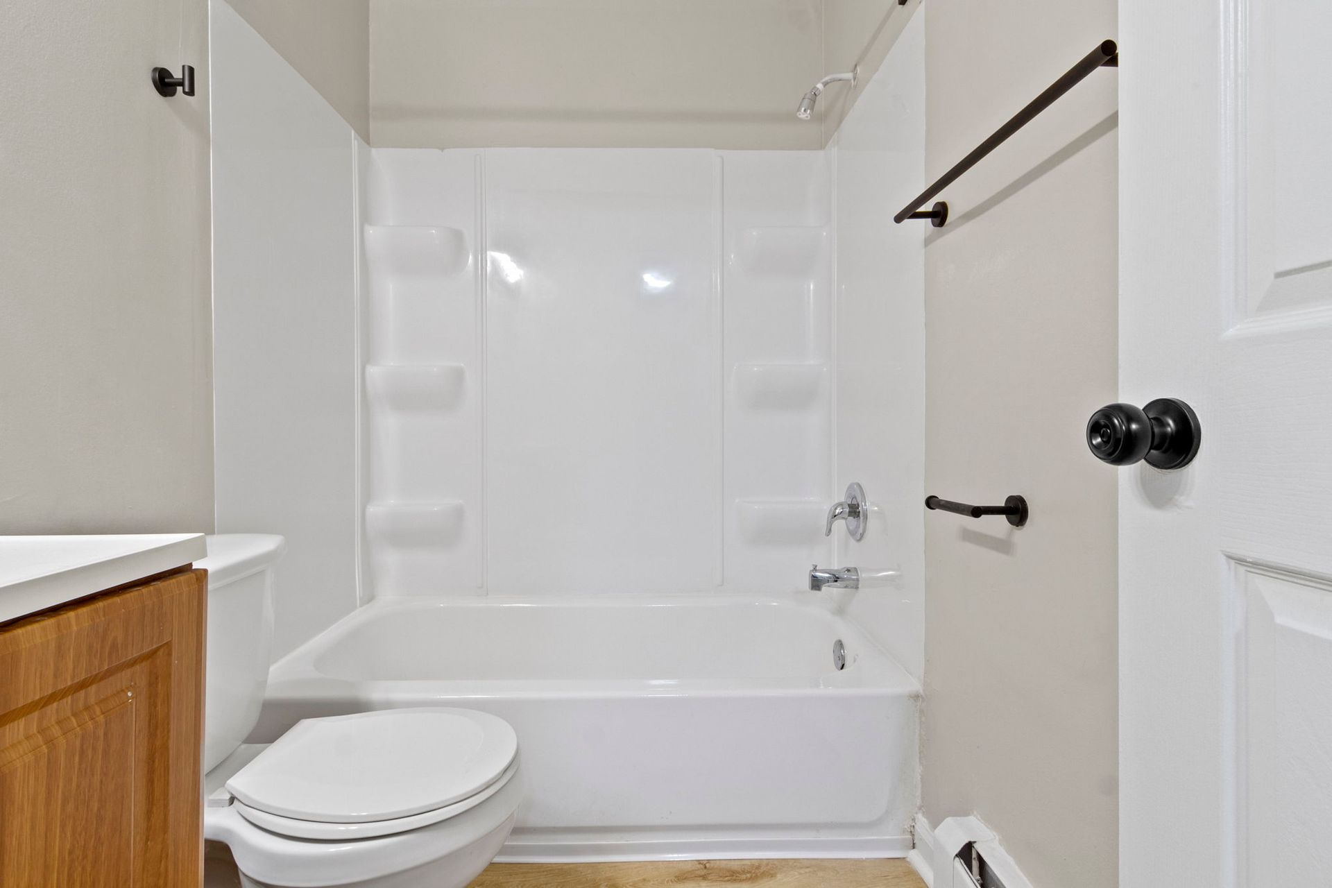 Small bathroom with white bathtub, toilet, and shower surround. Beige walls, brown cabinet, and black hardware.