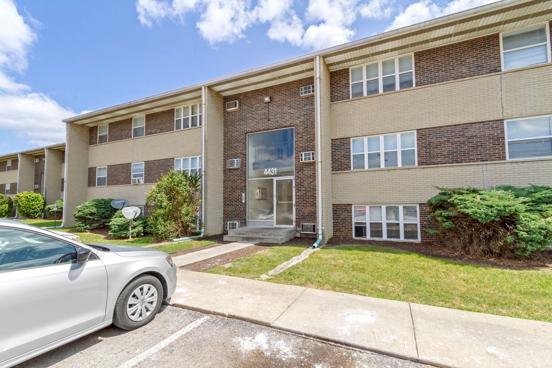 Two-story apartment building with brick and beige siding, a glass entryway, and a parked silver car.