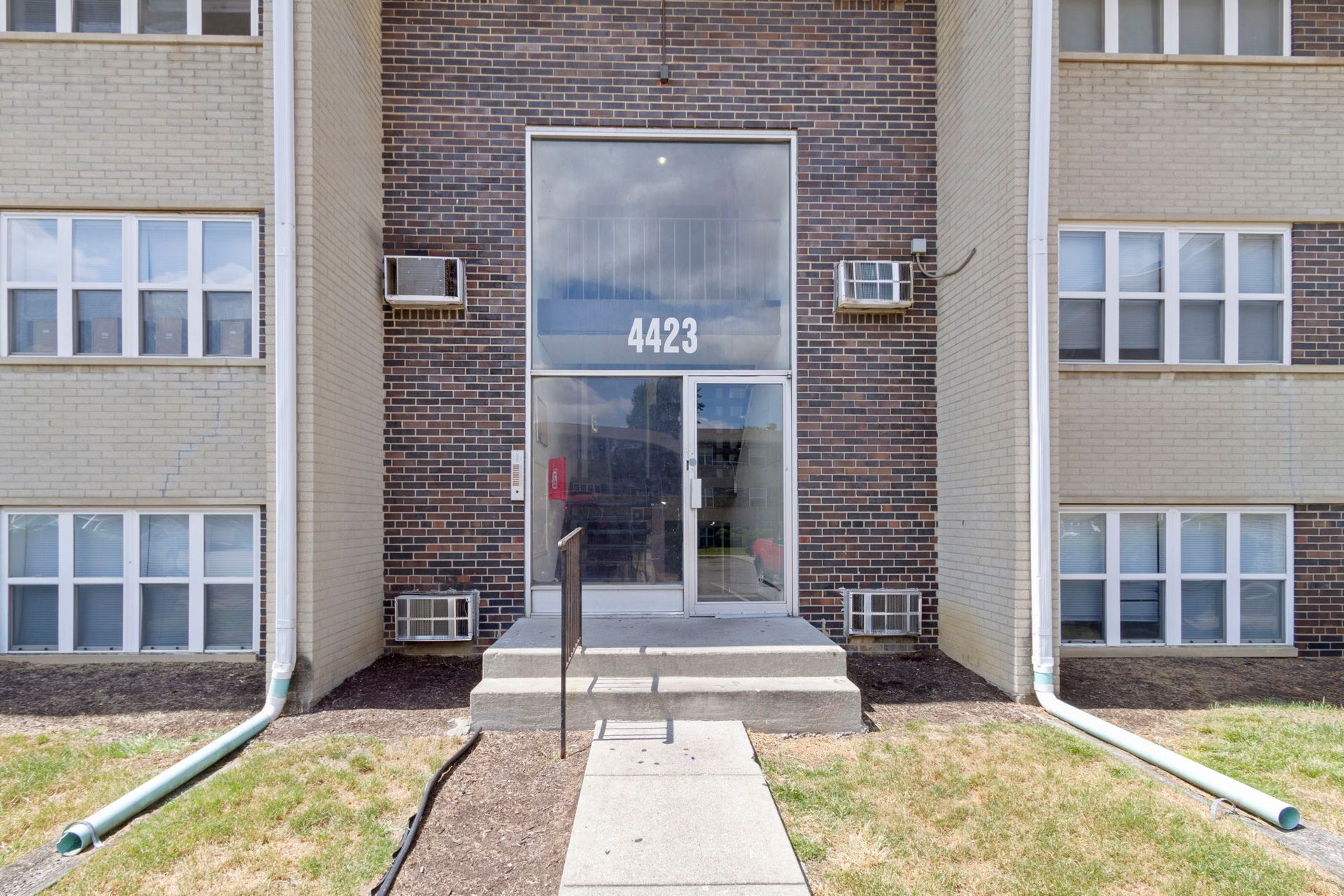 Exterior of apartment building with central entrance labeled 4423; brick and beige facade, air conditioners, and grassy lawn.