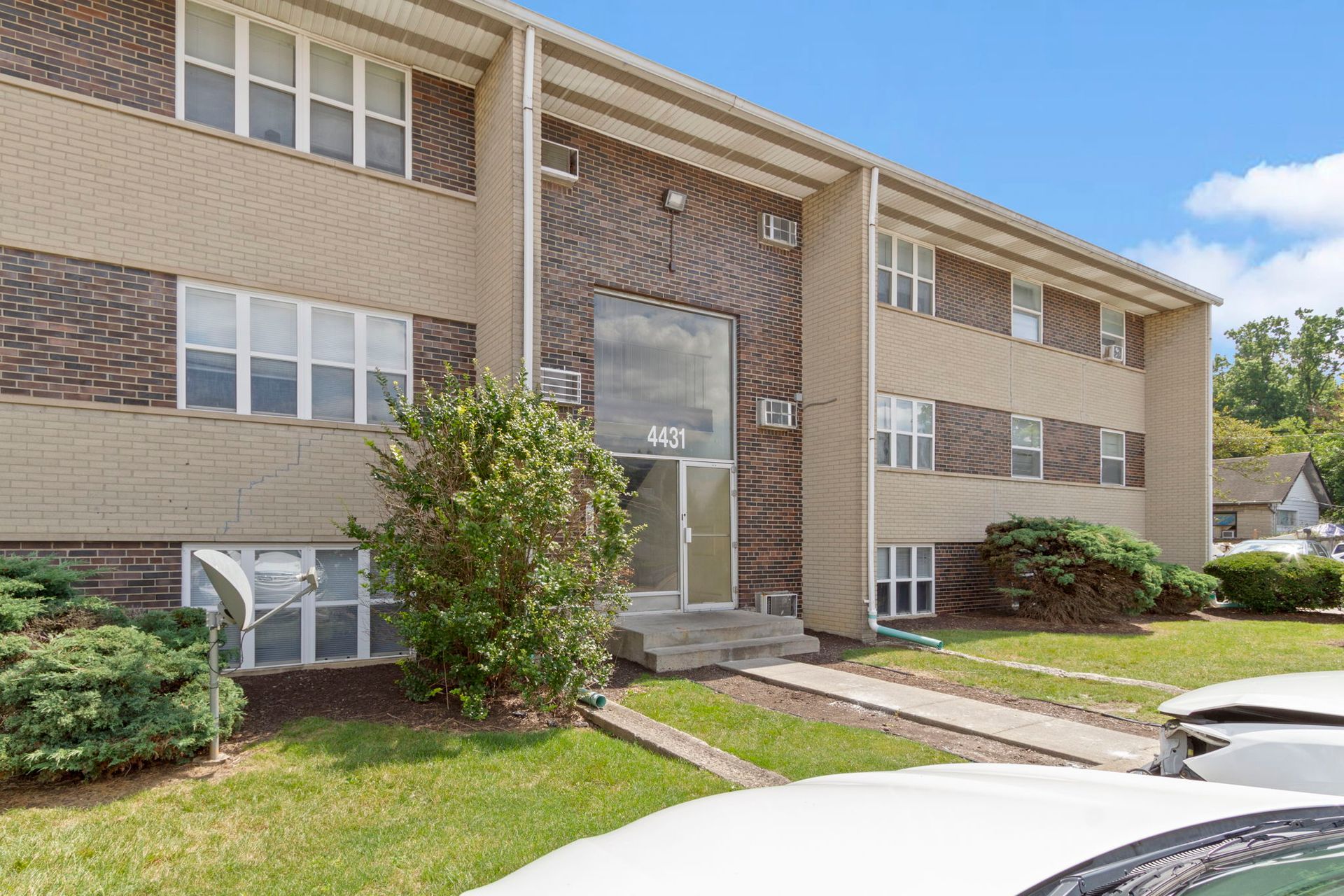 Two-story brick apartment building with a central glass entrance, green bushes, and parked cars.