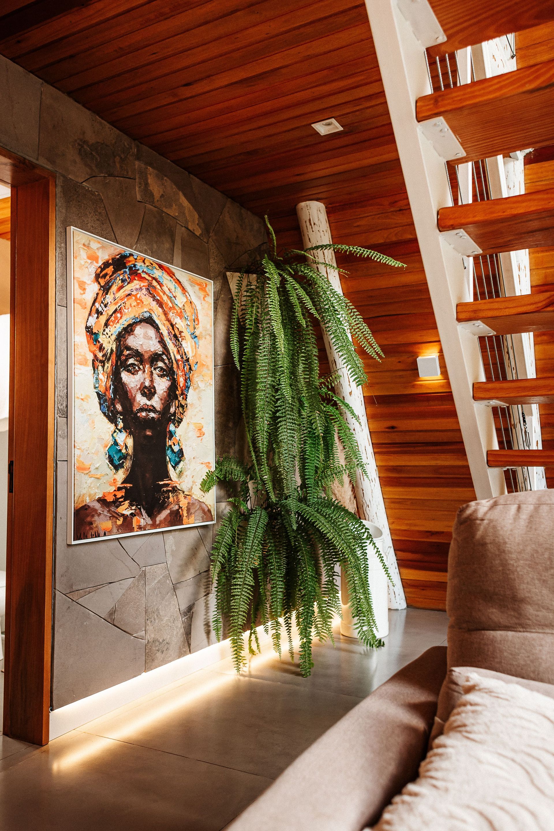 Corredor interno com obra de arte representando uma mulher usando um turbante, uma samambaia e escadas de madeira.