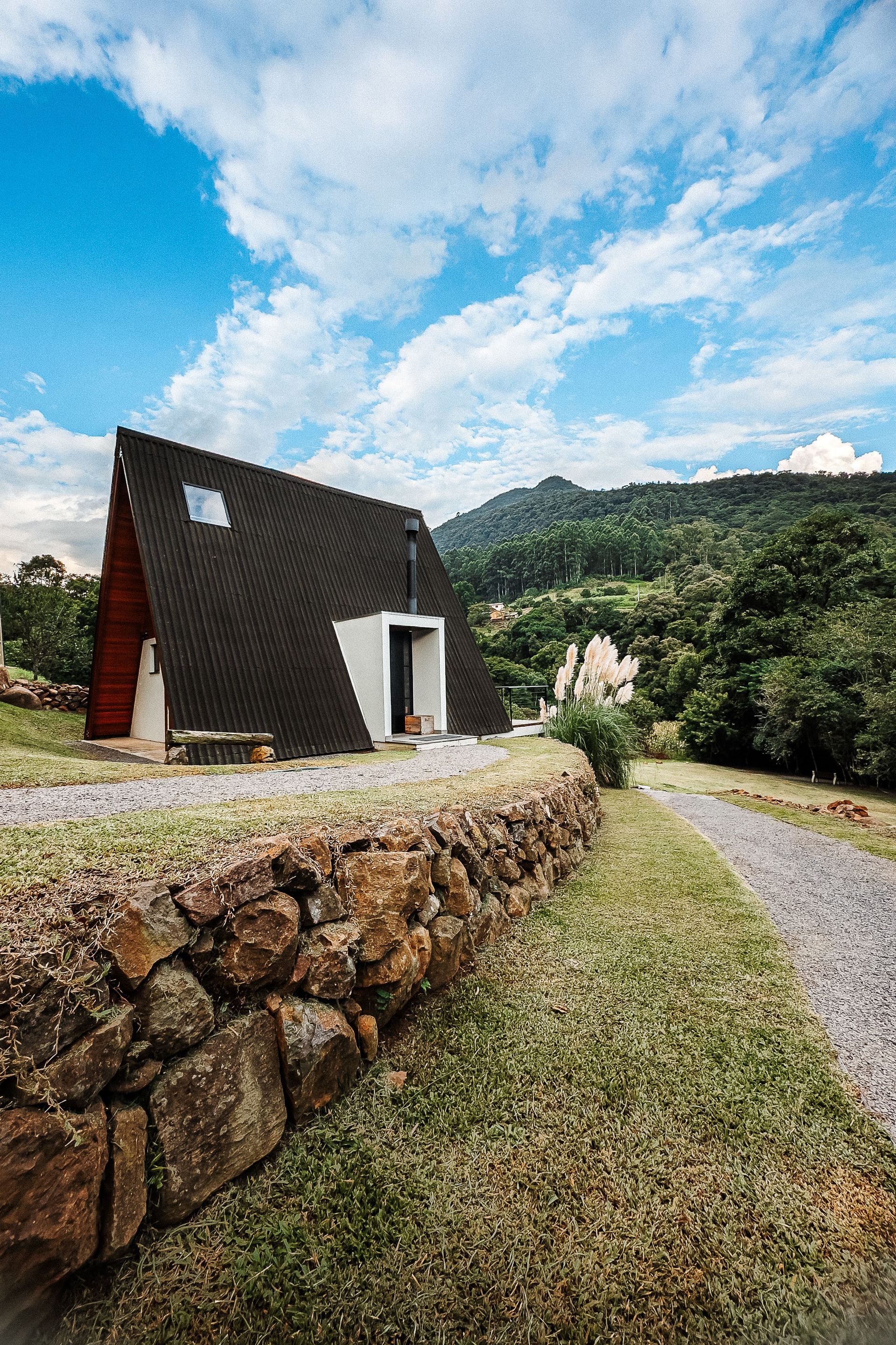 Cabana em forma de A com telhado escuro e detalhes brancos, situada em meio à grama verde e uma parede de pedra, sob um céu nublado.