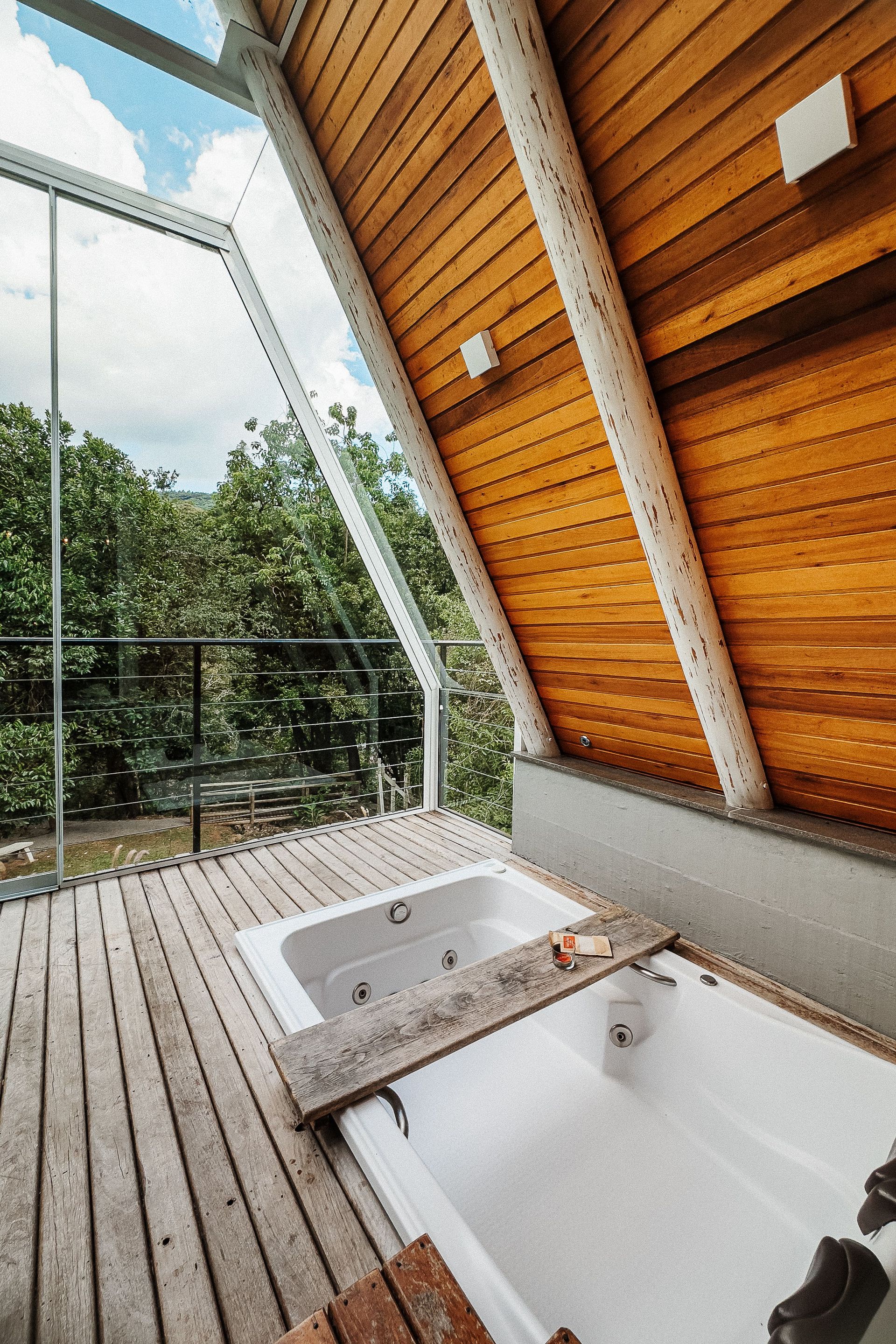 Duas banheiras brancas em um deck de madeira dentro de uma cabana com parede de vidro com vista para as árvores.
