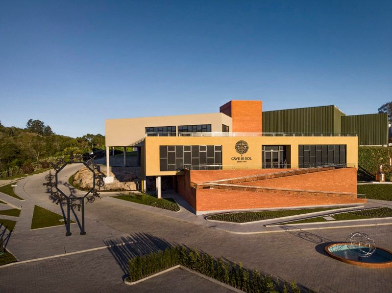 Prédio moderno com fachada de tijolos e tons de bege, emoldurado por um céu azul, com entrada pavimentada e área verde.