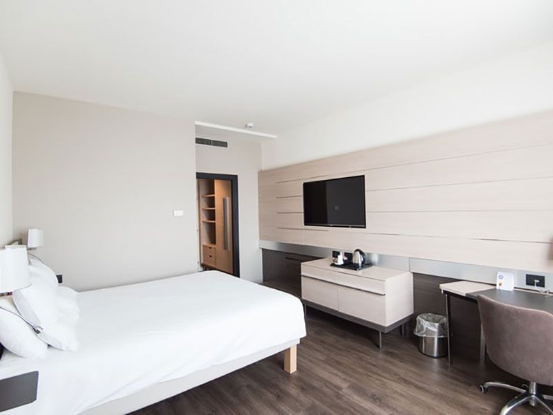 Quarto de hotel com cama, TV, escrivaninha e decoração em tons neutros.