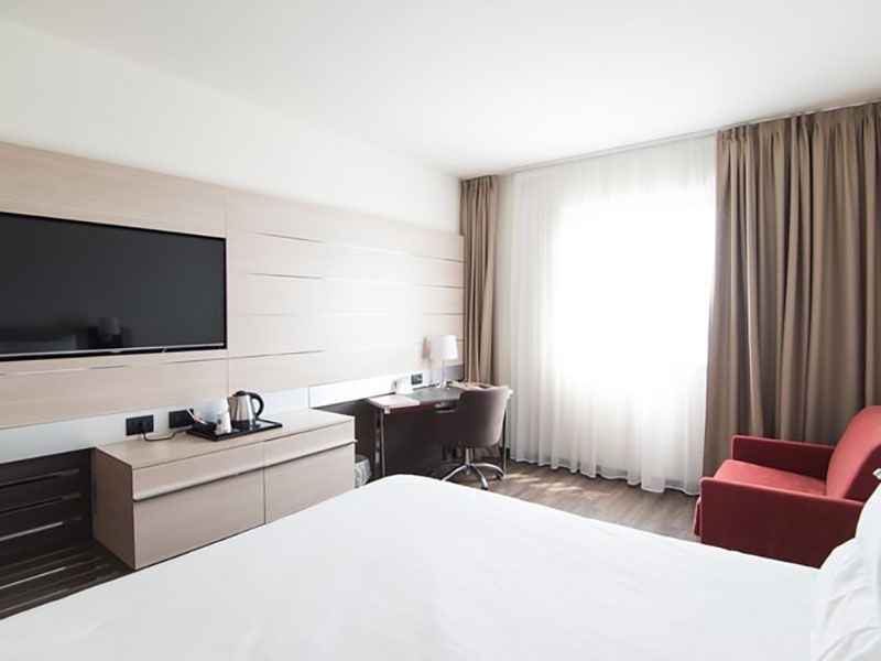 Quarto de hotel com cama, TV, escrivaninha, sofá pequeno e janela com cortinas.