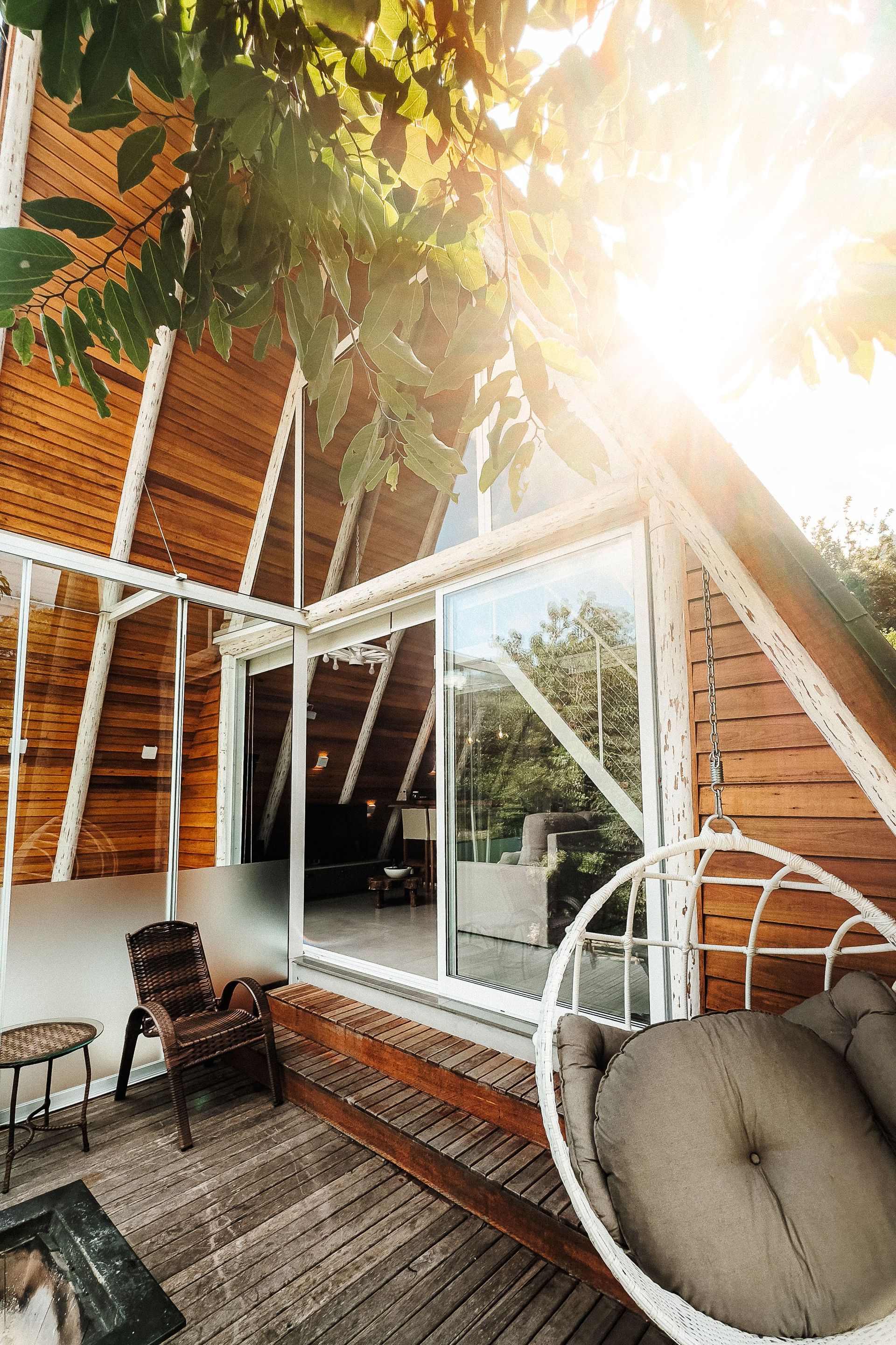 Cabana em forma de A com revestimento de madeira, grandes janelas de vidro e uma cadeira suspensa em um deck; sol brilhando através das árvores.