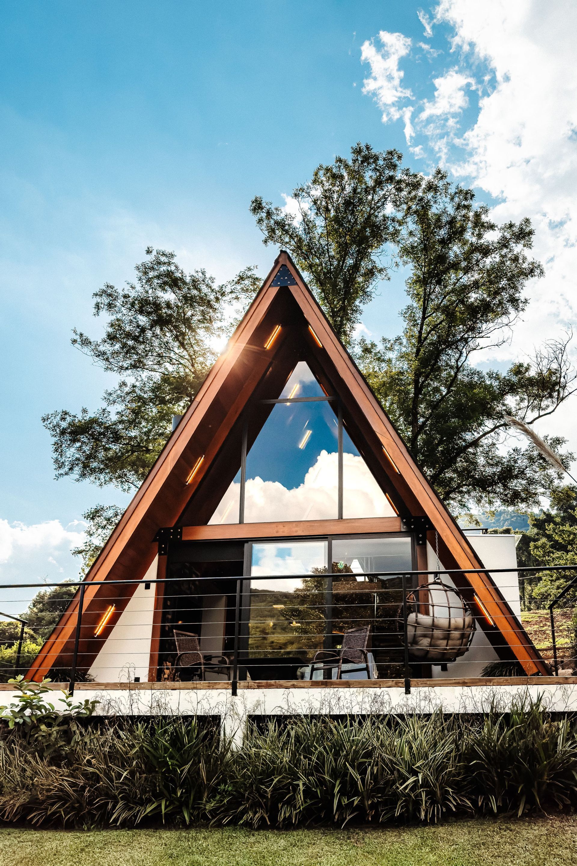 Cabana em forma de A com grande janela de vidro que reflete o céu, rodeada por árvores e vegetação.