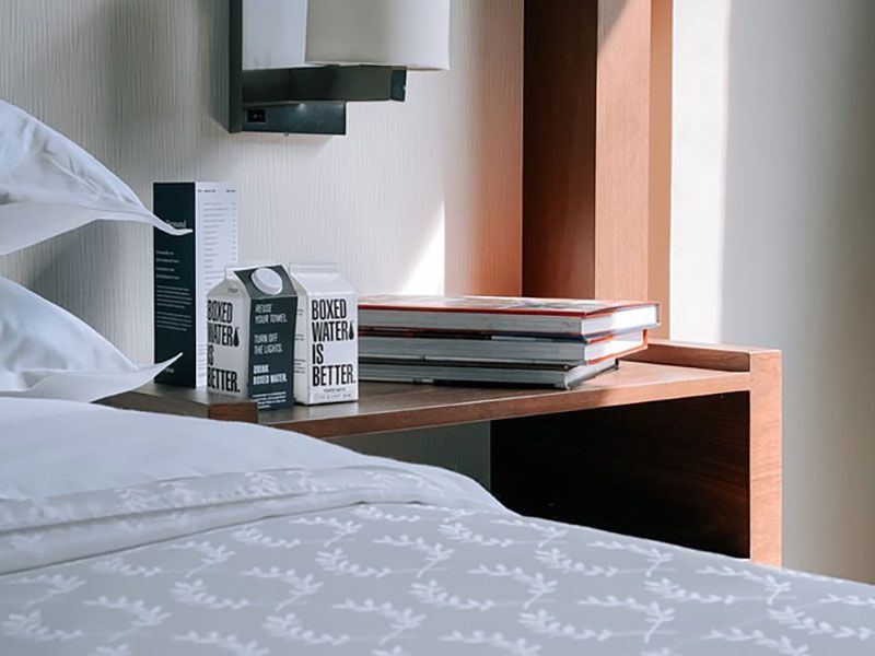 Mesa de cabeceira com pilha de livros, caixa de água, abajur e roupa de cama em um quarto.