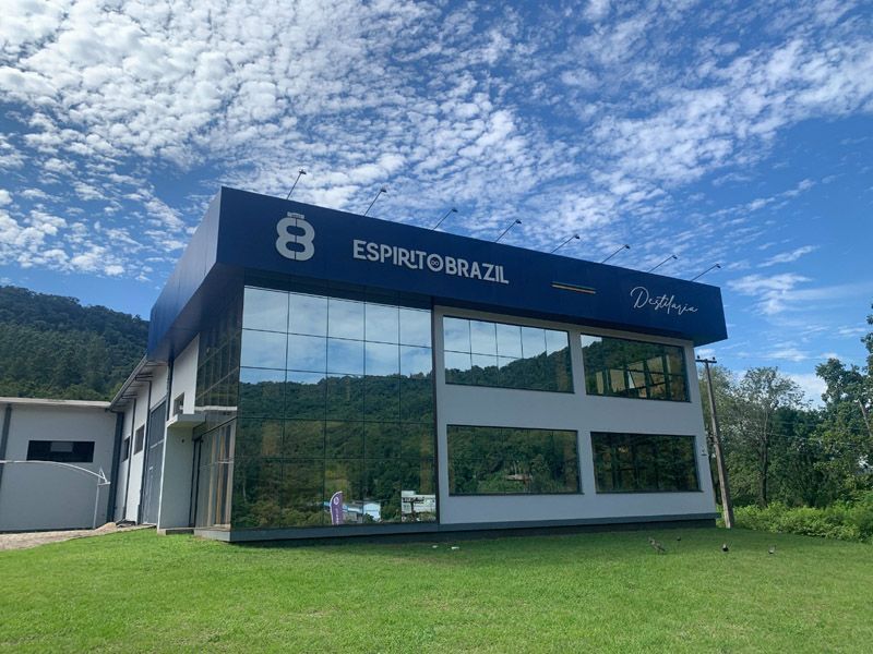 Prédio azul e branco com janelas espelhadas, placa "Espreto Brazil", em gramado verde sob céu azul.
