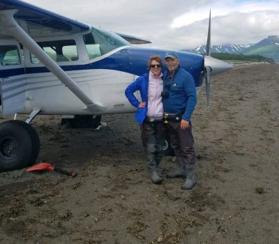 Two people pose with a small plane on a beach. Mountains in the background.