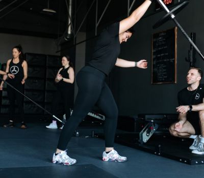 Woman lifts barbell in gym, assisted by trainer. Other people observe in the background.