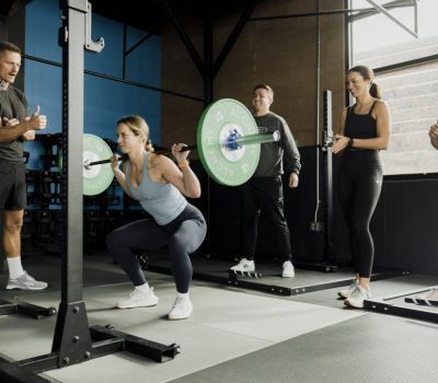 Woman doing squats with barbell, coached by others in a gym.