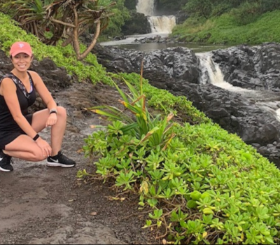 Woman squats on a path near waterfalls, wearing a pink cap and black athletic wear. Lush green vegetation surrounds the rocks.