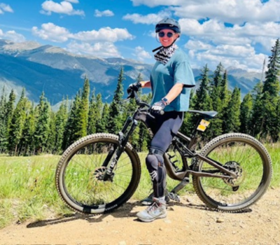 Woman on mountain bike, wearing helmet, gloves, and leg armor, on a dirt path with mountain backdrop.