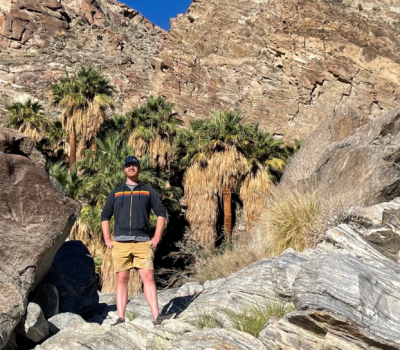 Man stands in rocky desert landscape with palm trees; he wears shorts, a jacket, and hat.
