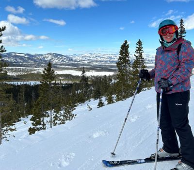 Person skiing on a snowy slope, holding poles, wearing a helmet and goggles, overlooking a valley.