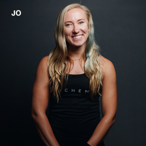 Jo smiles, wearing a black tank top. She has blonde hair, against a gray backdrop.