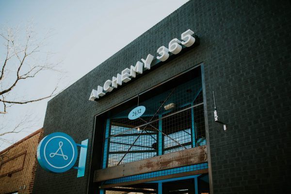Alchemy 365 storefront with a blue and white sign. Dark gray building with an open entrance.