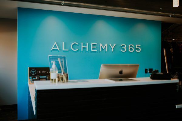 Reception desk for Alchemy 365 fitness studio. Turquoise wall with white logo, black counter, and Apple computer.