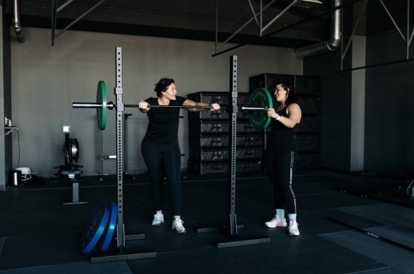 Two women spot a barbell at a gym; one adds a weight plate. Gray and black interior.