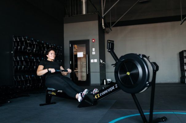 Woman rowing on a Concept2 machine in a gym, wearing black clothing, dark walls, and weights.