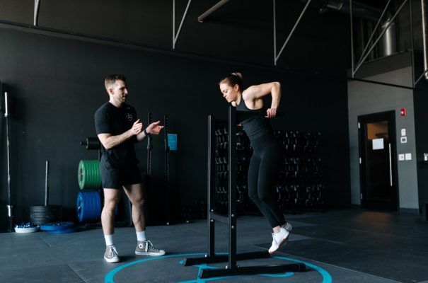 Man coaching woman doing dips in a gym. Woman is in the dip position with both arms extended and legs dangling.