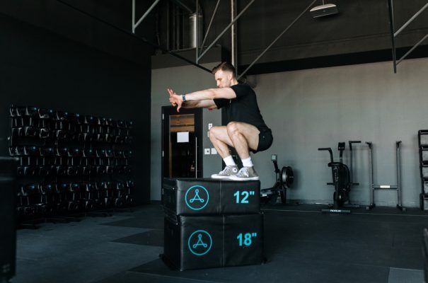Man performing a box jump in a gym. He is wearing black and gray athletic gear, squatting to jump on two boxes.