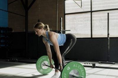 Woman in a gym, performing a deadlift exercise with a barbell. She's bending over, focused, with weights on the floor.