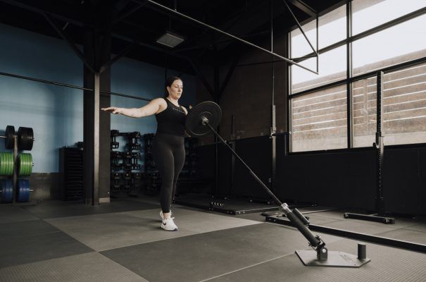 Woman using a landmine barbell in a gym, arms outstretched.
