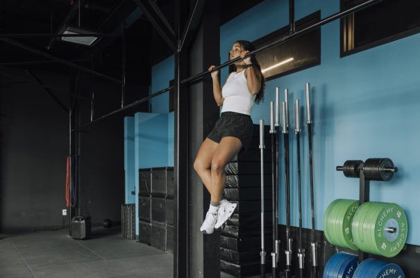 Woman doing pull-up in gym. Wearing shorts and tank top, dark hair, blue walls, weights and bars in the background.