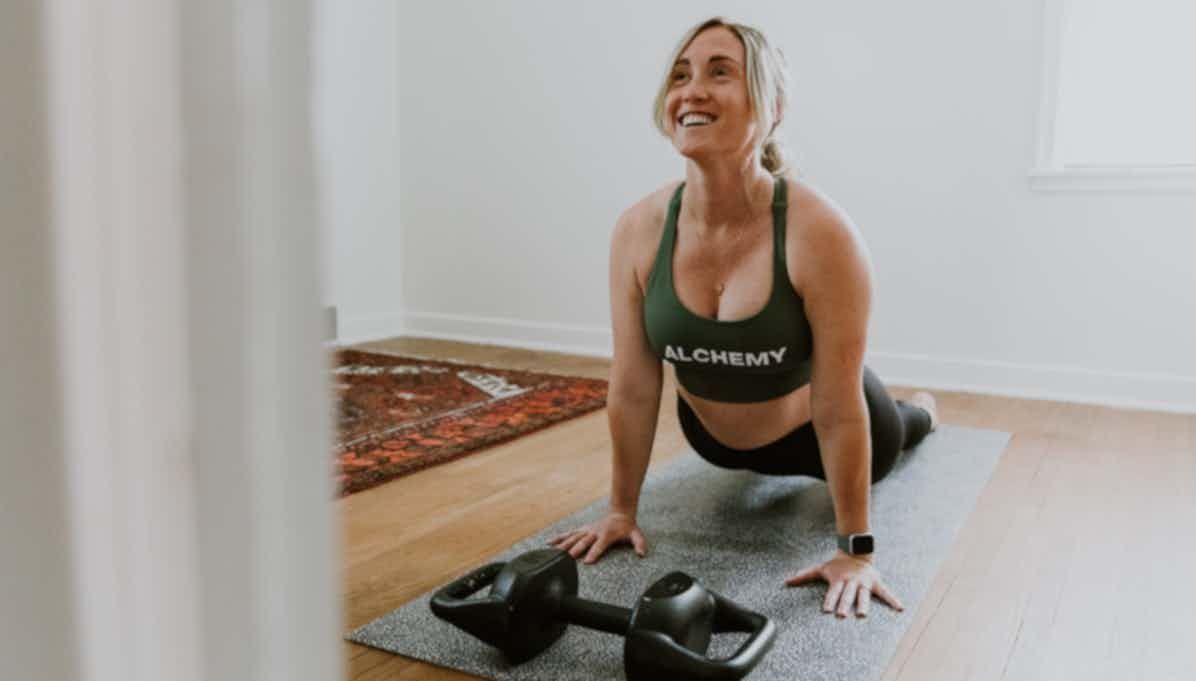 Woman in green sports bra doing a yoga pose, smiling. Black weight, gray mat, wooden floor in home.