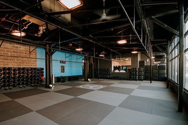 Spacious gym with dark floor and exposed beams; blue wall, weights, and windows on the right.