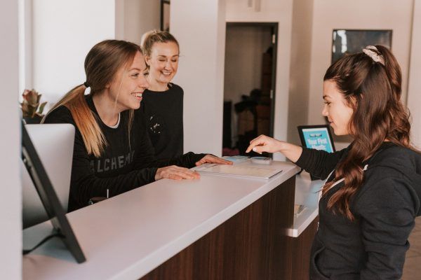 Three women at a reception desk: one pointing at a screen, two smiling. Neutral colors, indoor setting.