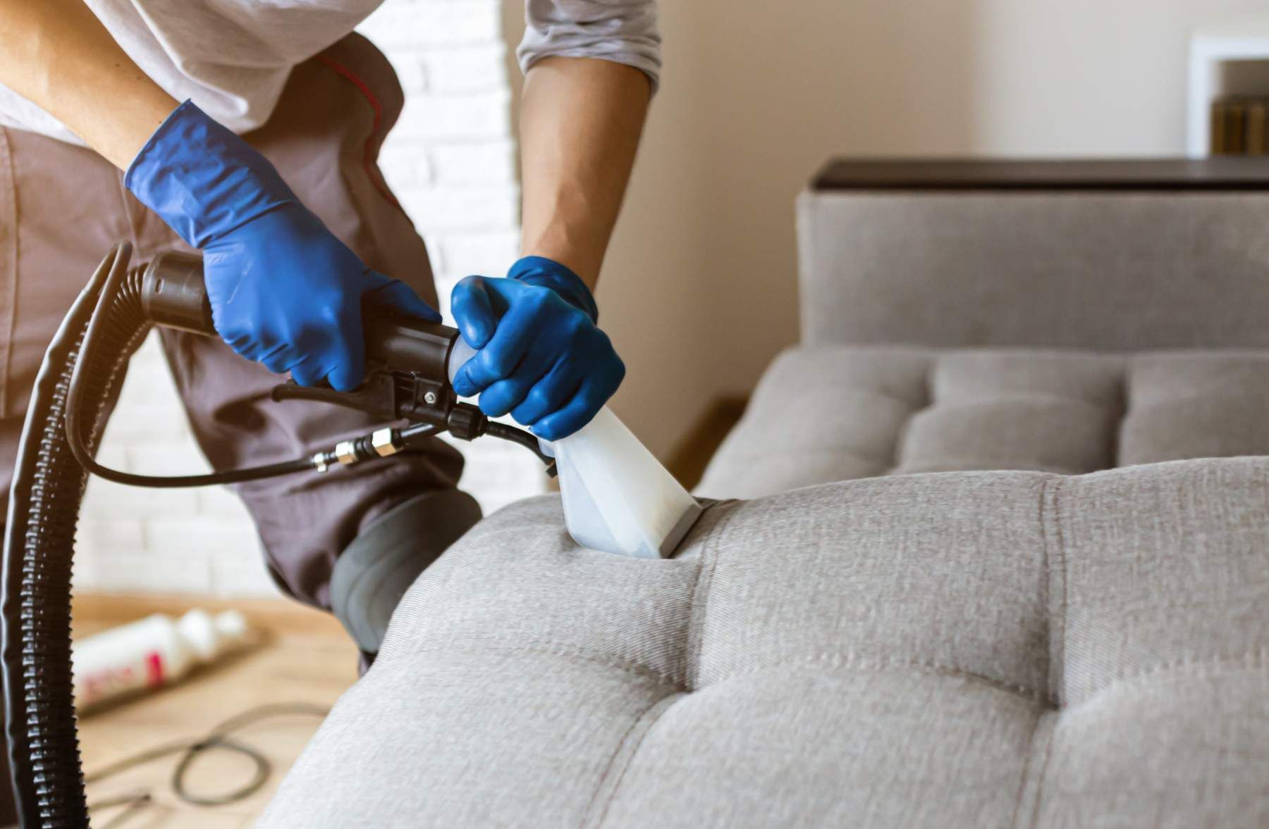 Professional Cleaner, cleaning a gray upholstered couch with a commercial steam cleaner.