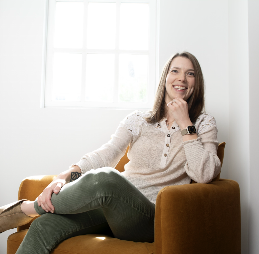 Sam with a slight smile sits smiling in a gold chair, angled toward the camera, with one leg crossed over the other.