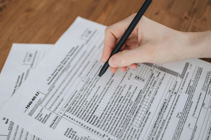 Hand holding a black pen writing on a stack of tax forms on a wooden desk.