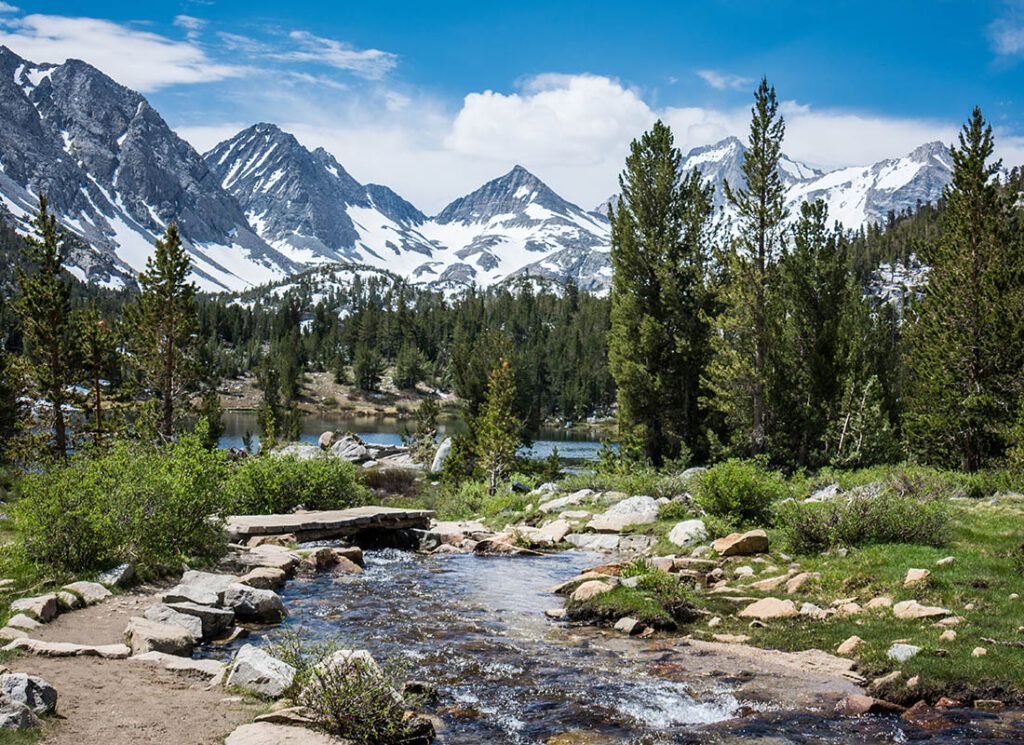 Mountain landscape with snow-capped peaks, pine trees, stream, and blue sky.