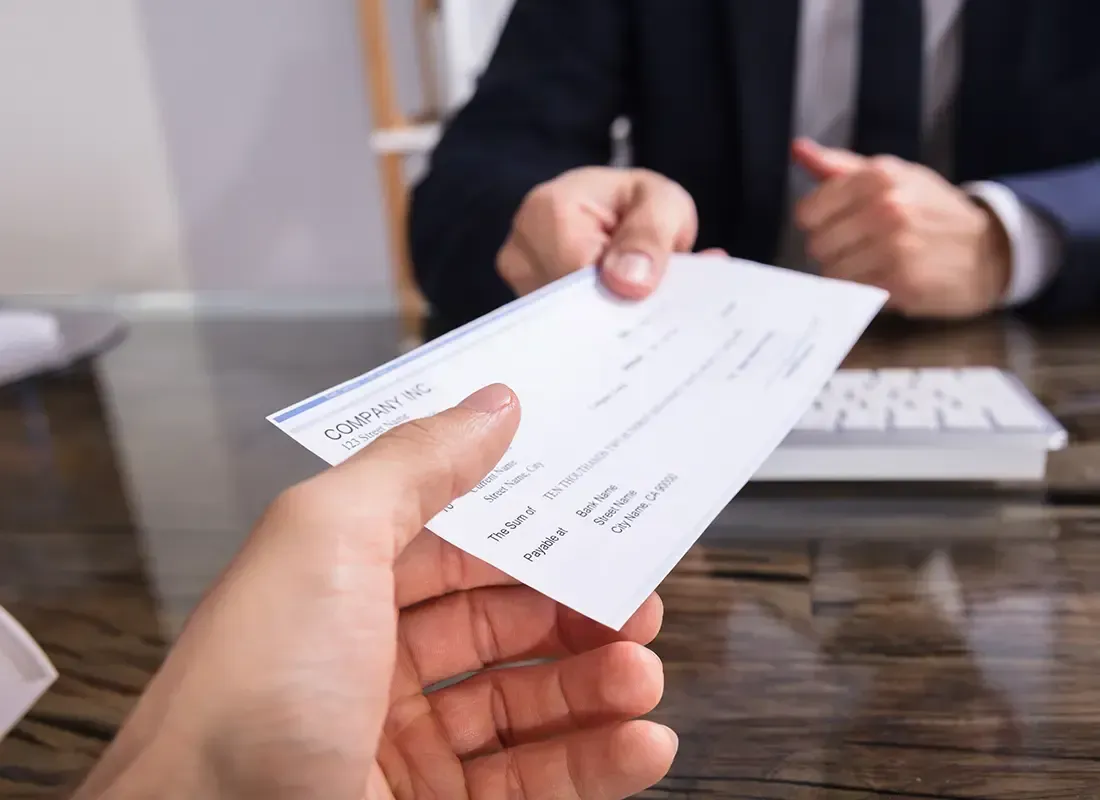 Hand receiving a check from another person at a desk in an office setting.