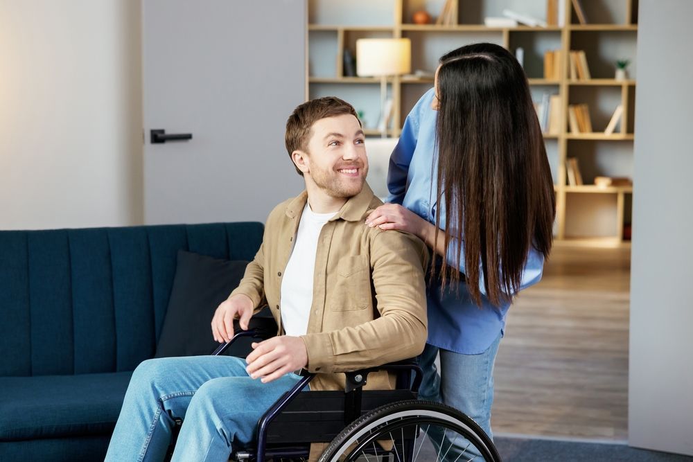 A Smiling Man In A Wheelchair Looks Up At A Woman Standing Behind Him — Comfort N Care Support QLD Pty Ltd In Pittsworth, QLD
