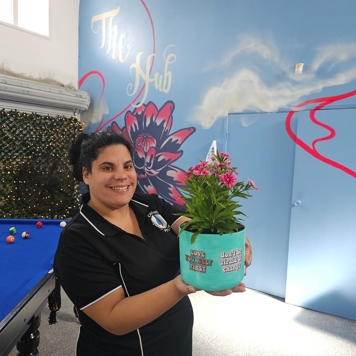 A Woman Is Holding a Potted Plant in Front of A Pool Table that Says the Hub — Comfort N Care Support QLD Pty Ltd In Highfields, QLD