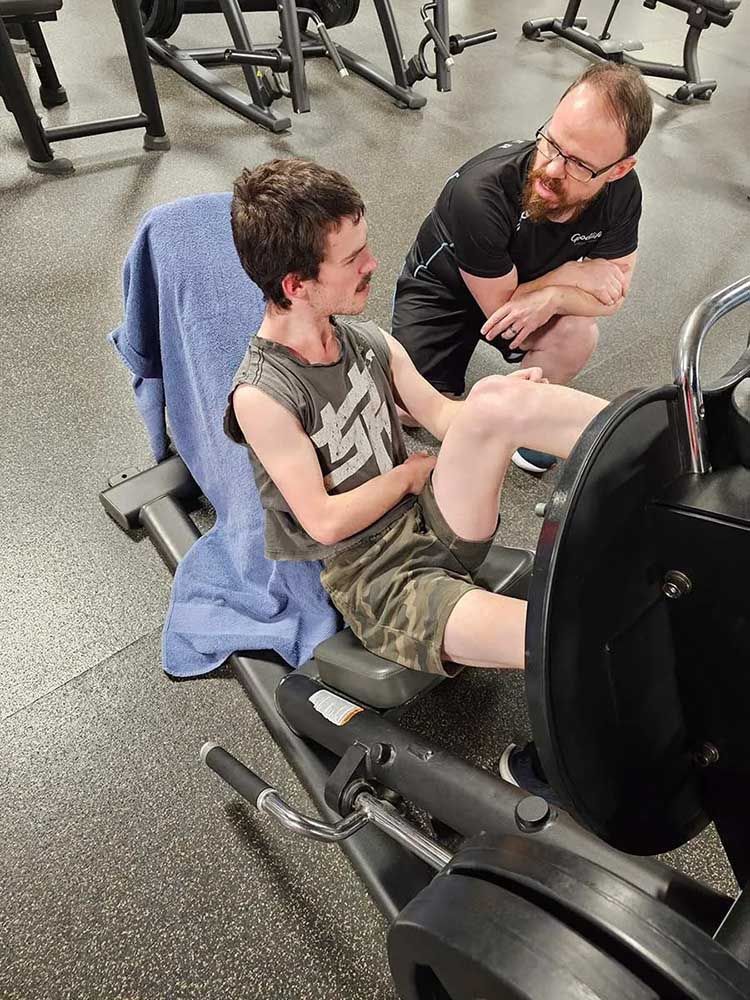 A Man Is Talking to A Man Sitting on A Machine in A Gym — Comfort N Care Support QLD Pty Ltd In Toowoomba City, QLD
