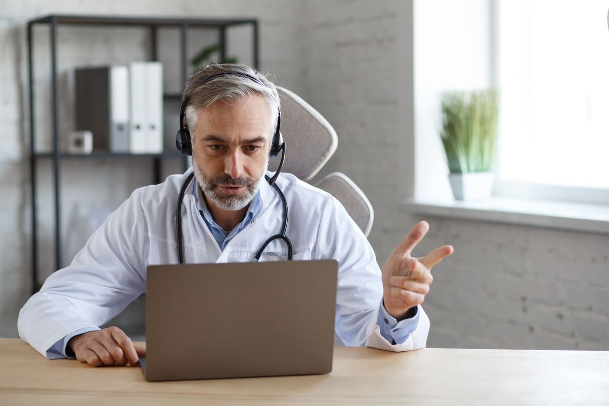 Um médico usando fones de ouvido está sentado em uma mesa em frente a um laptop.