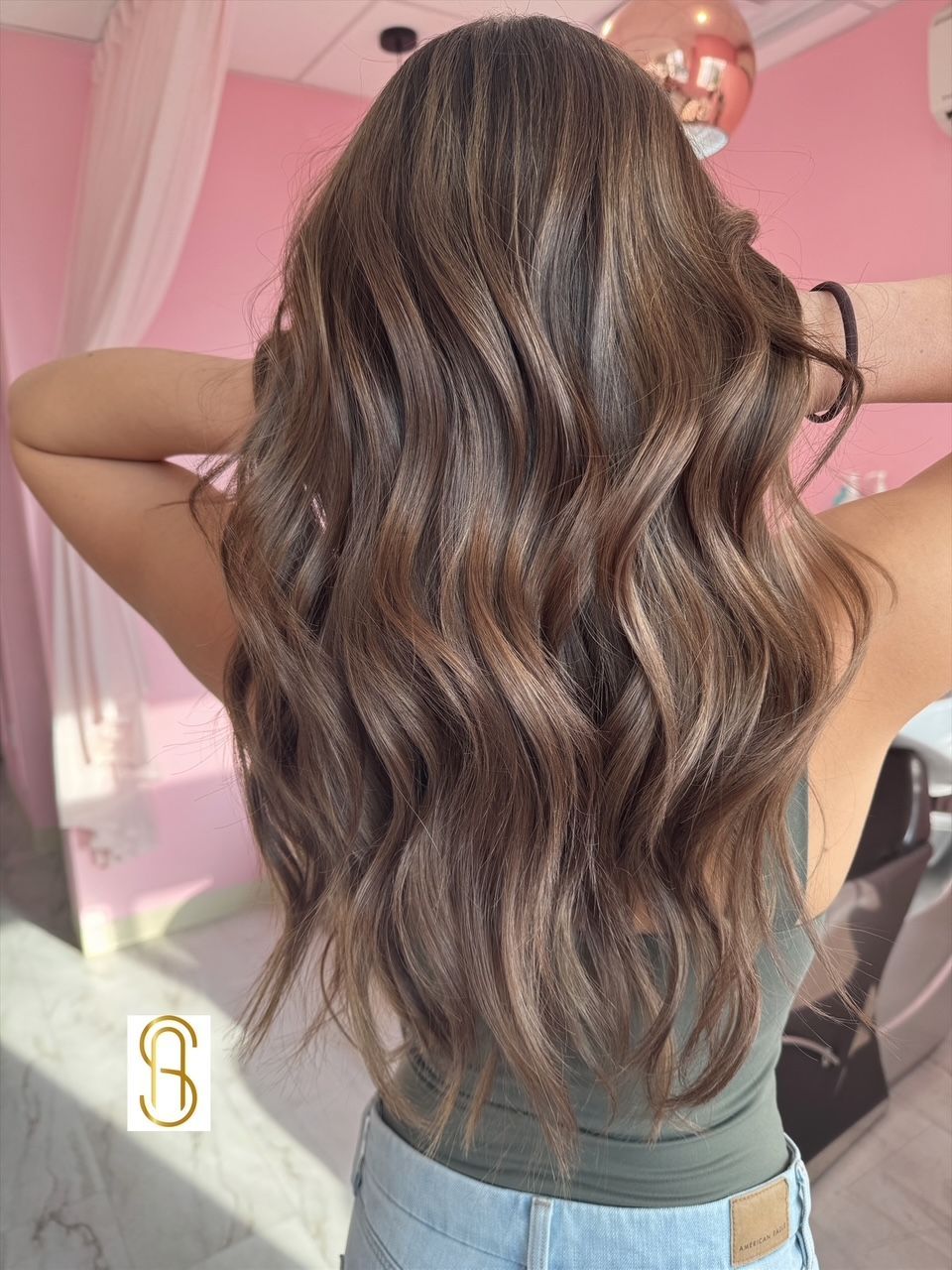 A woman with long wavy brown hair is standing in front of a pink wall.
